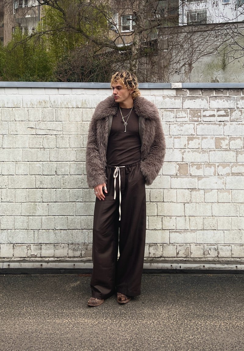 Personne aux cheveux blonds bouclés portant une veste en fausse fourrure marron, un pantalon large marron foncé avec un cordon blanc, et des colliers superposés, debout en plein air.