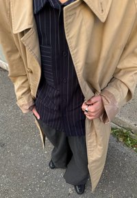 Trench coat bege com um design de duplo peito, usado sobre uma camisa riscada azul-marinho. Calças cinzas de perna larga e sapatos pretos completam o visual.