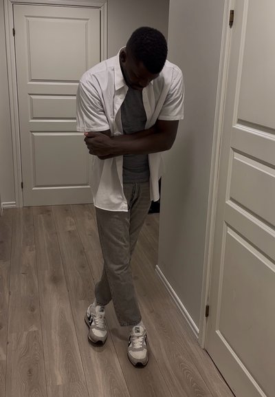 Hombre con los brazos y las piernas cruzados está de pie en un pasillo vistiendo una camisa blanca, camiseta gris, pantalones grises y zapatillas blancas. Cabeza inclinada hacia abajo.