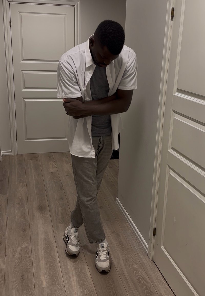 Homme aux bras et jambes croisés se tenant dans un couloir, portant une chemise blanche, un t-shirt gris, un pantalon gris et des baskets blanches. Tête baissée.