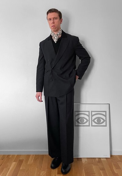 Hombre con traje negro cruzado, zapatos negros y una bufanda blanca con estampado, de pie junto a una pared blanca y al lado de una obra de arte enmarcada.