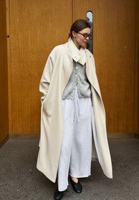 Femme portant un manteau crème oversize, un cardigan gris, un pantalon blanc large, des chaussures noires et des lunettes, debout devant des portes en bois.