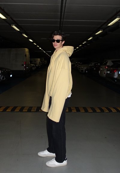 Sudadera amarilla de gran tamaño con cordón, pantalones negros y zapatillas blancas. El fondo presenta un garaje de estacionamiento con poca luz y coches aparcados.