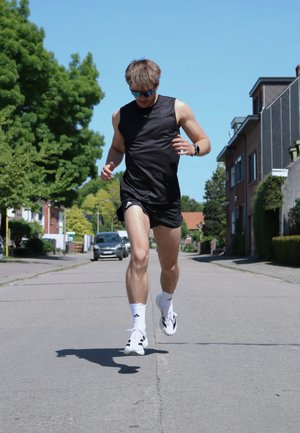 Scarpe da corsa indossate con calze bianche, canottiera nera, pantaloni corti neri e occhiali da sole; corridore su una strada fiancheggiata da alberi e case.