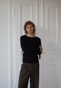 Camicia nera a maniche lunghe con scollo rotondo e bottoni, abbinata a pantaloni marroni a righe sottili. Sullo sfondo pareti bianche a pannelli.