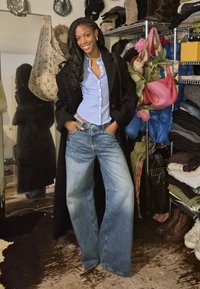 Jean bleu clair à jambes larges, une chemise bleu rayée ajustée et un manteau noir, debout dans une pièce remplie de sacs et de vêtements.