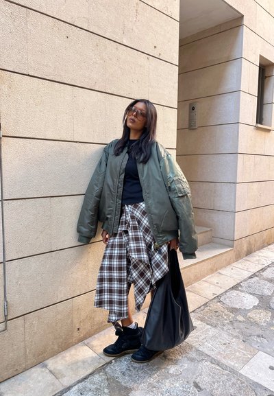 Chaqueta bomber verde, camiseta negra, falda envolvente de cuadros, zapatillas negras y un gran bolso negro. El conjunto presenta una variedad de texturas y patrones.