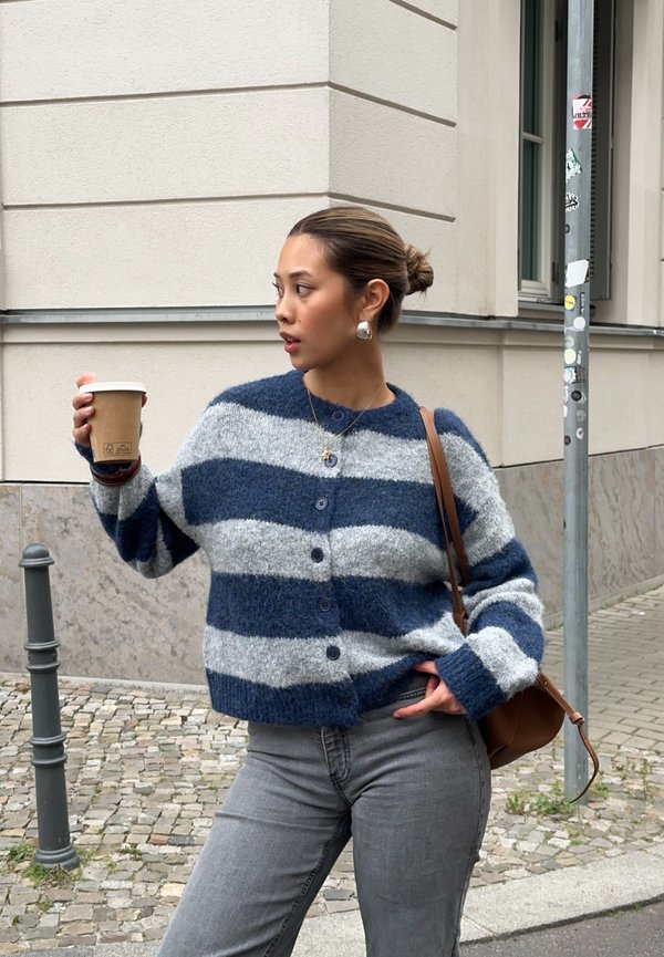 Blauwe en grijze gestreepte cardigan van een zachte brei, met knopen aan de voorkant en een ontspannen pasvorm, gecombineerd met grijze jeans. Koffiekop in de hand.