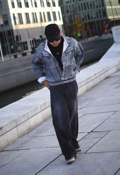 Chaqueta de mezclilla con cuello blanco, camiseta negra, vaqueros oscuros holgados y gorra negra. Escenario al aire libre con edificios modernos de fondo.