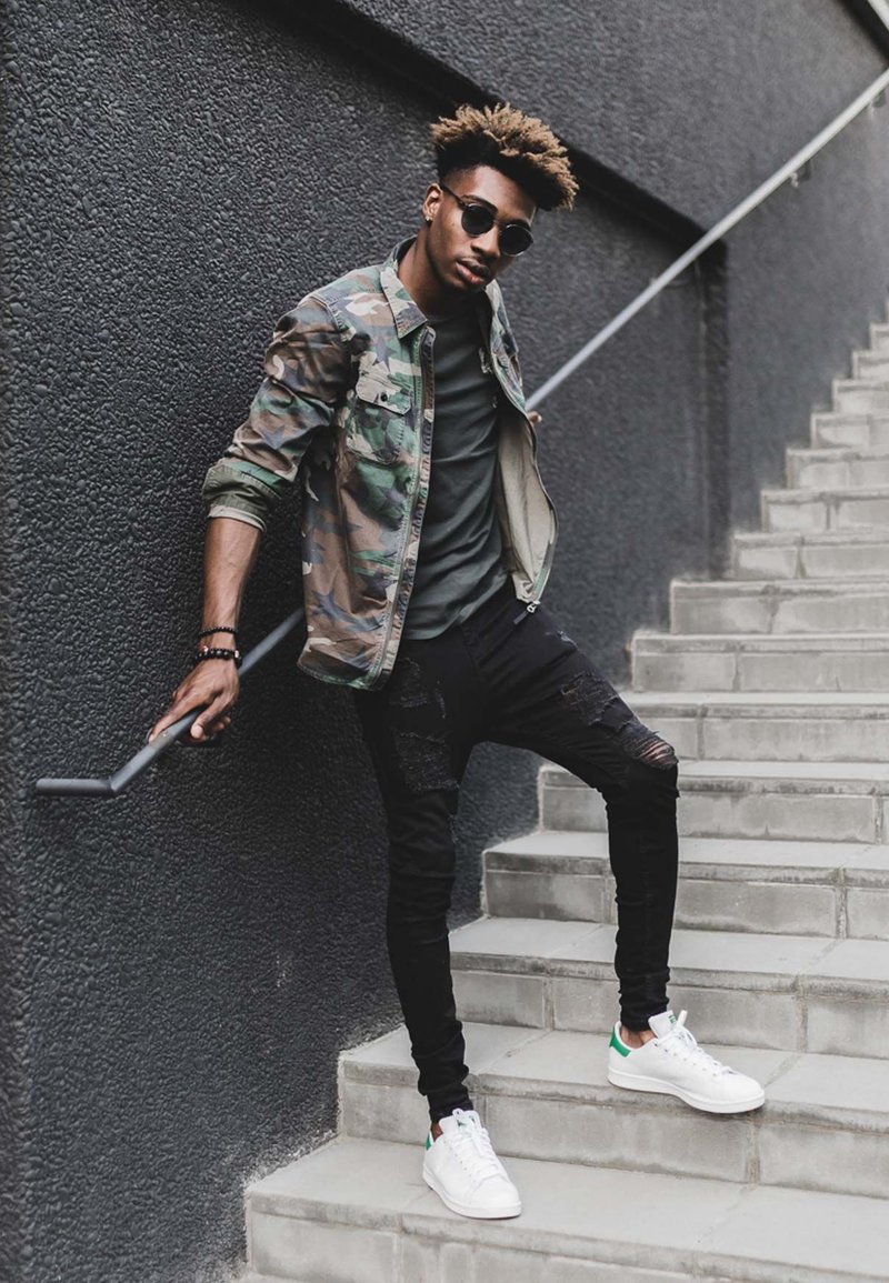 Camouflage jacket over a grey shirt, black ripped skinny jeans, and white sneakers with green accents, standing on stairs against a dark wall.