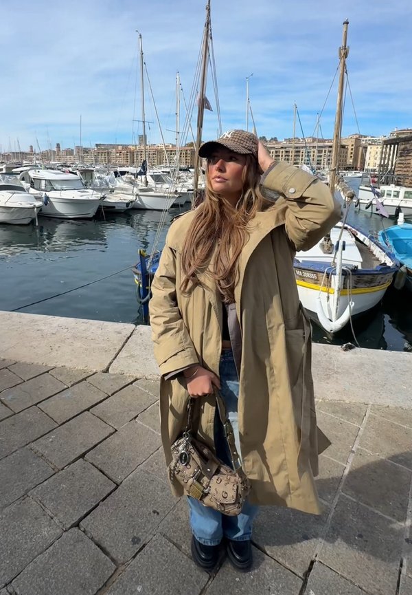 Une femme en manteau beige et casquette pose à côté de voiliers amarrés dans une marina ensoleillée, en tenant un sac à main à motifs.