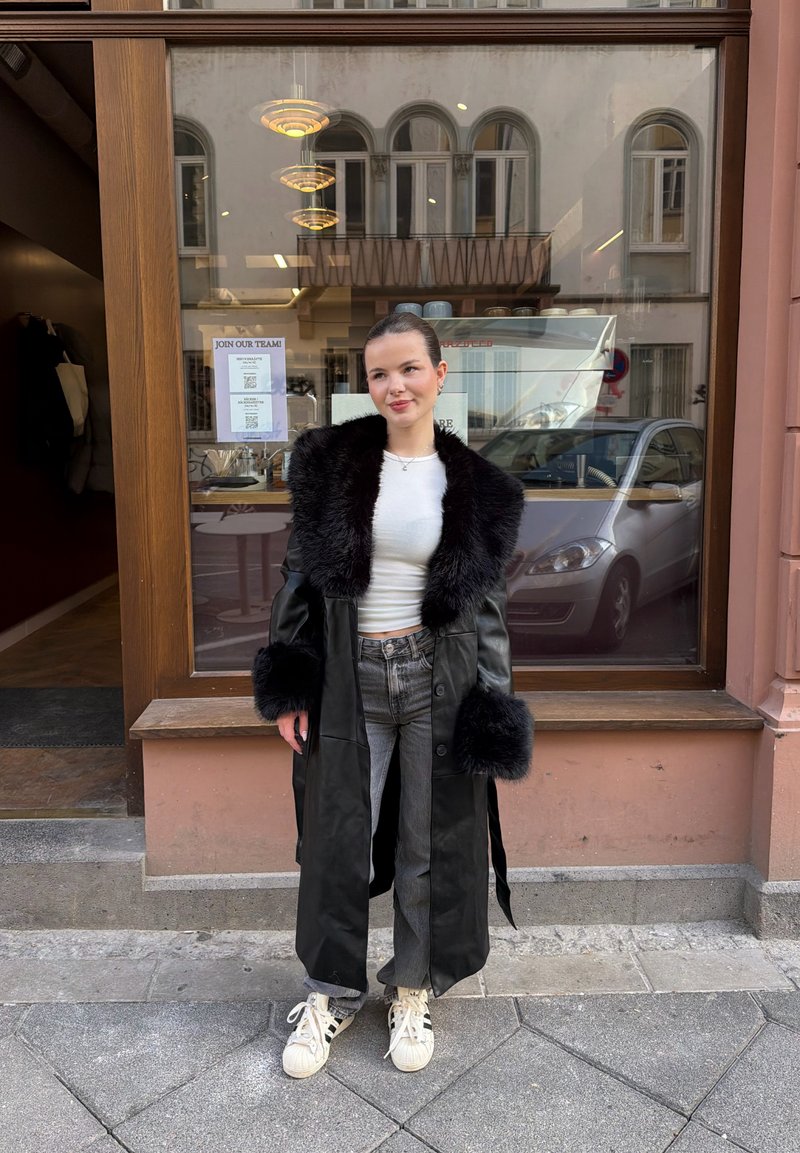 Jauna sieviete melnā mētelī ar kažokādas apmali, baltā krekliņā, pelēnās džinsās un sporta apavos stāv ārpus kafejnīcas ar lielu logu un tajā atspoguļotām noliktajām automašīnām.