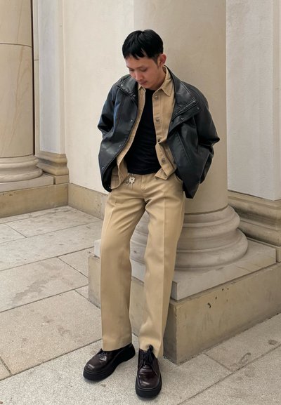 Chaqueta de cuero negra sobre una camisa beige de botones, camiseta negra, pantalones beiges y zapatos de plataforma marrón oscuro. Columnas en el fondo.