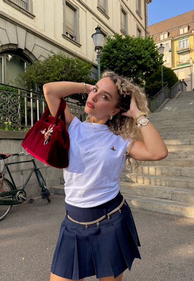 Camisa blanca de manga corta con detalle bordado, falda plisada azul marino con acentos de cadena y un bolso de terciopelo rojo con charms decorativos.