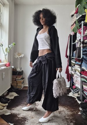 Mujer con peinado afro posando en interior, vestida con camiseta sin mangas blanca, chaqueta negra, pantalones anchos negros, tacones blancos y sosteniendo un bolso blanco con flecos.
