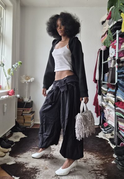 Mujer con peinado afro posando en interior, vestida con camiseta sin mangas blanca, chaqueta negra, pantalones anchos negros, tacones blancos y sosteniendo un bolso blanco con flecos.