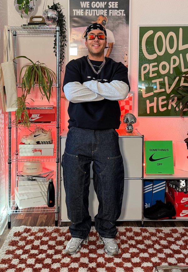 Homme portant des lunettes de soleil, un bandana, un collier en chaîne, un sweat-shirt et un jean, les bras croisés dans une pièce colorée avec des chaussures, des plantes et des affiches.