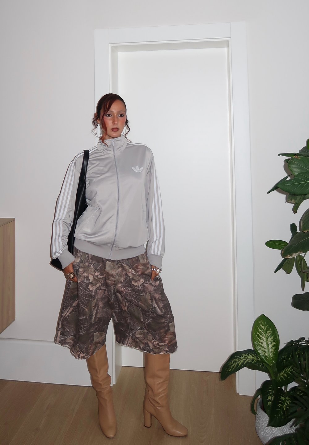 Henkilö, joka on pukeutunut vaaleanharmaaseen Adidas-urheilu takkiin, kamouflagekuvioisiin leveisiin shortseihin, tanvärisiin korkokenkiin ja kantaa mustaa olkalaukkua sisätiloissa oven ja kasvien vieressä.