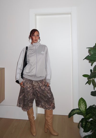 Persona vestida con chaqueta de chándal Adidas gris claro, pantalones cortos anchos de patrón de camuflaje, botas de tacón marrones, y llevando un bolso negro de hombro dentro de un edificio, junto a una puerta y plantas.