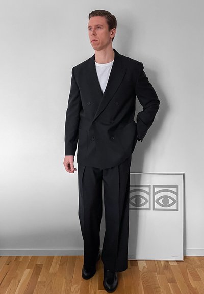 Hombre con traje negro cruzado y camiseta blanca de pie sobre suelo de madera junto a una pared con una obra enmarcada que muestra dos ojos abstractos.