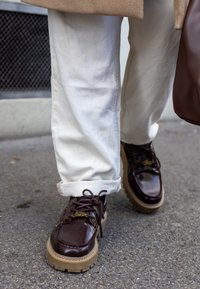 Hnedé lakované kožené topánky s textúrovanou svetlohnedou podrážkou, s tkaničkami a logom. Nosia sa s vyhrnutými svetlými nohavicami.