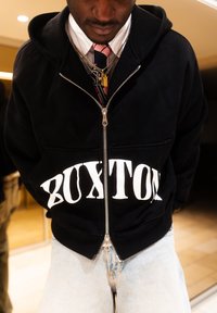 Άνδρας που φοράει μαύρη ζακέτα με φερμουάρ και την επιγραφή «BUIXTON», ροζ και μαύρη γραβάτα, πολλαπλά χρυσά κολιέ και ανοιχτόχρωμα μπλε τζιν.