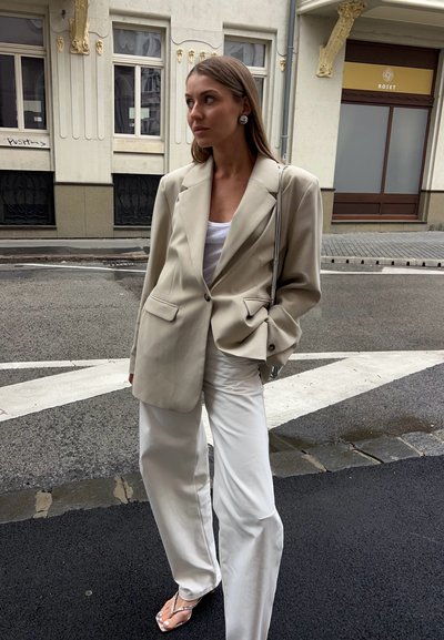 Blazer beige de gran tamaño con solapas notch y bolsillos frontales, combinado con pantalones de pierna ancha blancos y sandalias transparentes de tacón.