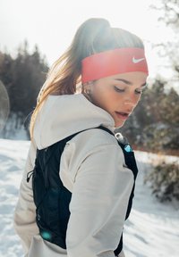 Jeune femme en tenue de sport d'hiver avec un bandeau rouge Nike regarde vers le bas, debout en plein air dans une forêt enneigée avec le soleil derrière elle.