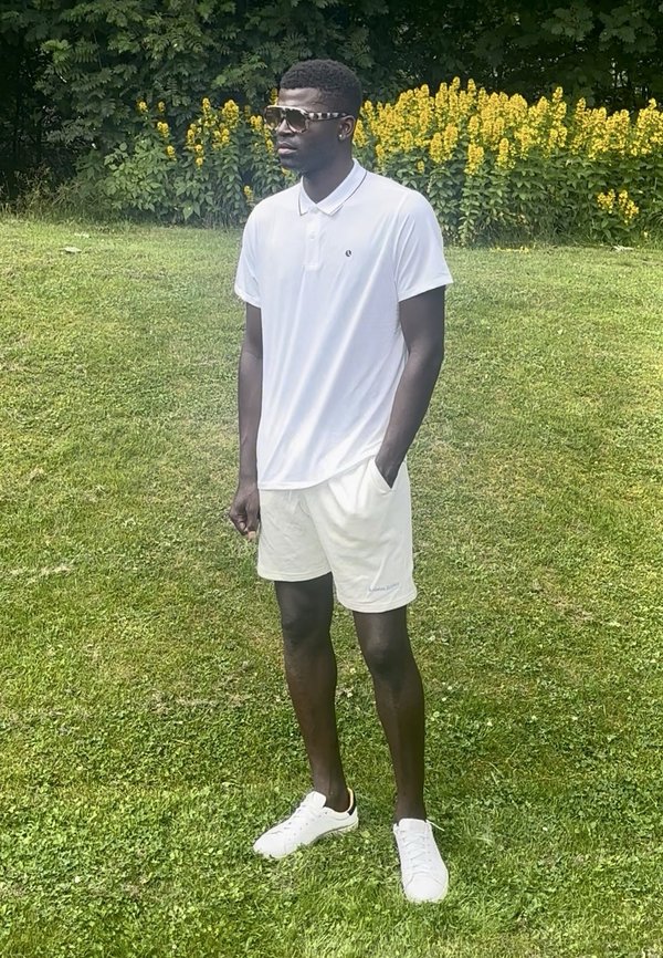 Man met een wit poloshirt, witte korte broek en witte sneakers staat op gras met gele bloemen en groene struiken achter zich.