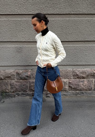 Cárdigan de punto trenzado crema, suéter negro de cuello alto, jeans acampanados azules, bolso de hombro de cuero marrón y botas de tacón de ante marrón. Fondo texturizado.