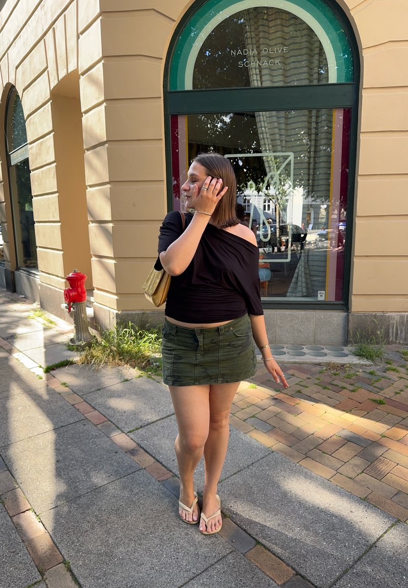 Young woman standing on a sunlit sidewalk, wearing a black top, green skirt, and flip-flops, with a beige shoulder bag.