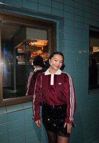 Top de satén burdeos con cuello blanco, mangas con tres rayas y logo bordado, combinado con falda negra de lentejuelas, frente a una pared de azulejos.