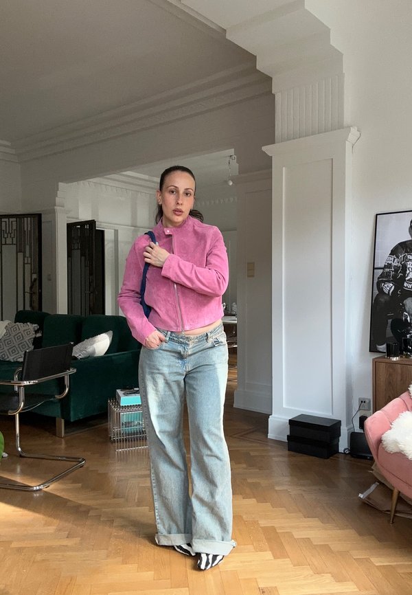 Femme debout à l’intérieur portant une veste rose, un jean bleu clair et des chaussettes à motif zèbre dans un salon spacieux avec un sol en bois.