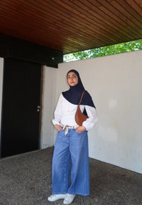 Weißes Langarmoberteil, weit geschnittene blaue Jeans, braune Handtasche, weiße Sneakers und ein dunkelblauer Hijab. Steht vor einer Betonwand.