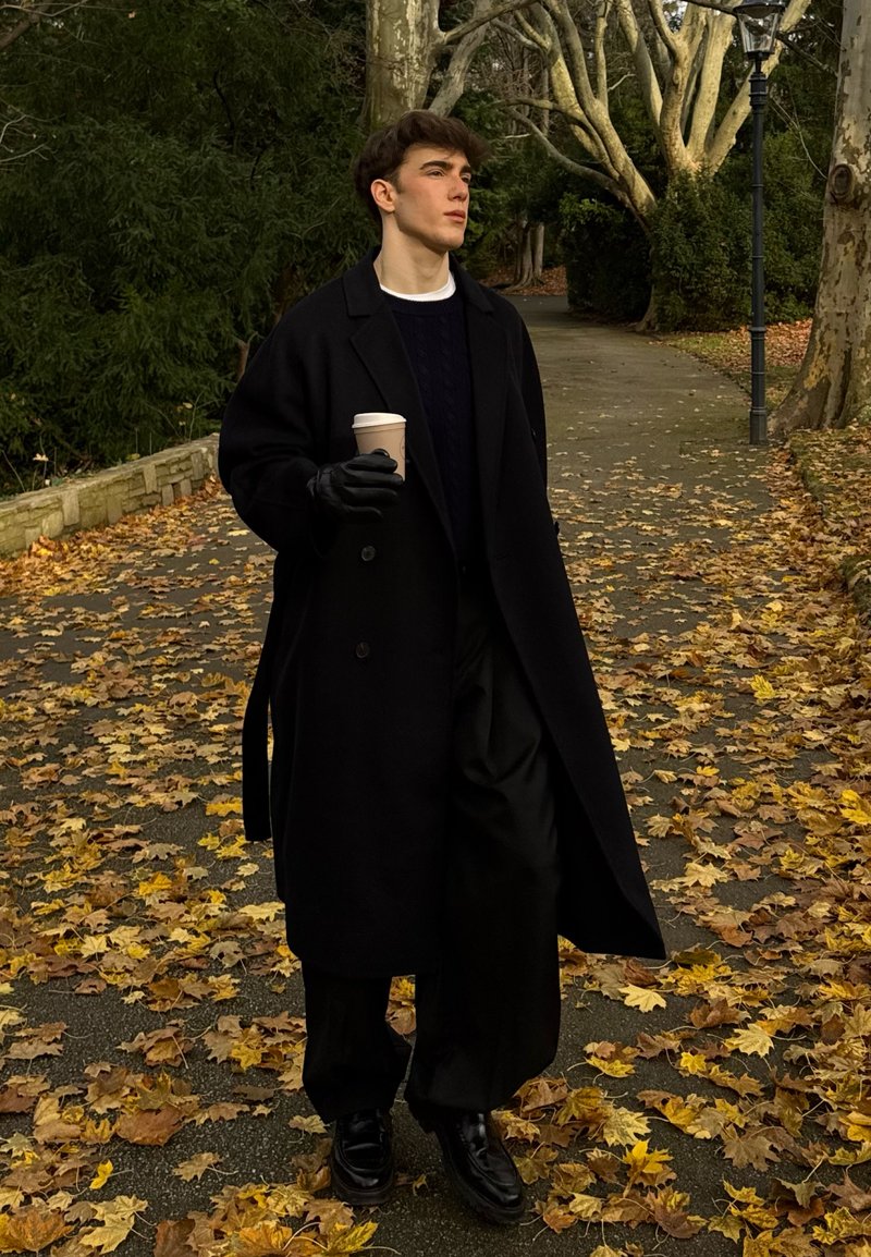 Manteau noir par-dessus un pull bleu marine, pantalon large noir et gants. Tenant un gobelet à café jetable, debout sur un chemin couvert de feuilles d'automne.