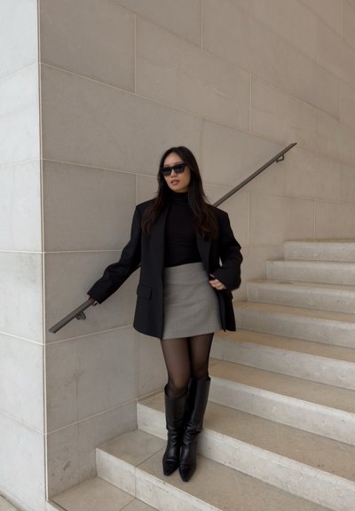 Blazer negro de gran tamaño con un top de cuello alto, falda corta de pata de gallo, medias negras transparentes y botas negras hasta la rodilla en una escalera.
