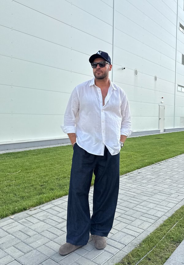 Bela prugasta srajca s gumbi, ohlapne temne denim hlače, sive semiš čevlje, črna kapa in sončna očala. Stoji na tlakovanem hodniku.