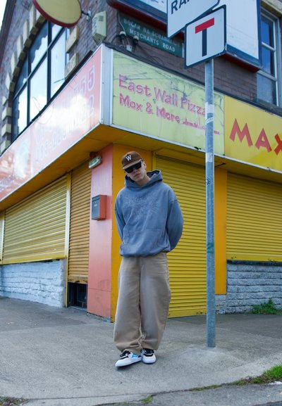 Sudadera gris con capucha, pantalones anchos beige y zapatillas negras con acentos azules. El fondo presenta una tienda cerrada con persianas amarillas.