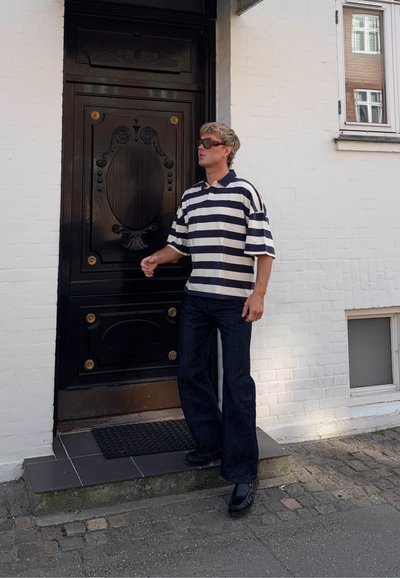 Hombre con camisa de rayas y gafas de sol de pie en la puerta delante de una puerta negra ornamentada en un edificio de ladrillo blanco.