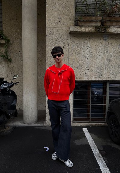 Joven con gafas de sol negras, suéter rojo con cuello atado, pantalones anchos oscuros y zapatos sin cordones a cuadros, de pie en una calle urbana.