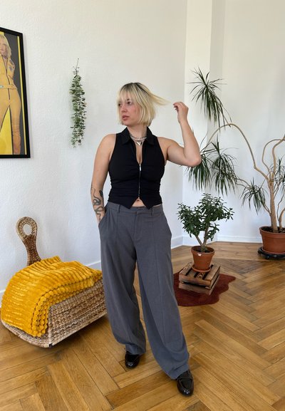 Top negro sin mangas con cremallera y diseño ajustado, combinado con pantalones grises sueltos. El fondo incluye plantas y una cesta tejida con una manta amarilla.