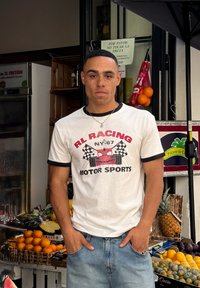 Bela majica z črnimi robovi, ki ima grafiko rdečega avtomobila in napis "RL RACING NY-67 MOTOR SPORTS." Oblečena je z modrimi džinsom; v ozadju je stojalo s sadjem.