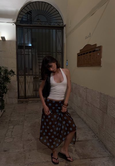 Top halter blanco, falda marrón con lunares azules, usando chanclas, de pie en un corredor de piedra con una puerta de metal al fondo.