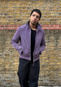 Violetti bomber-takki vetoketjulla ja sivutaskuilla, päällä musta paita. Takki näyttää sileältä tiiliseinää vasten.