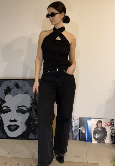 Mujer con top negro de tirantes y vaqueros de pierna ancha, con gafas de sol, sosteniendo un bolso, de pie en interiores cerca de una obra de arte de Marilyn Monroe y discos de vinilo.