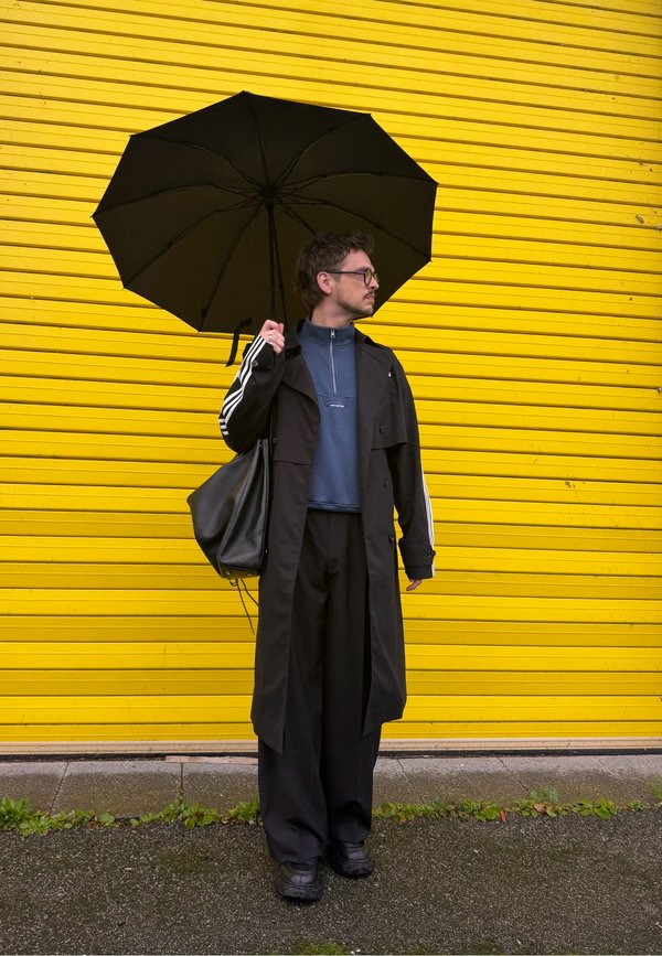 Un homme vêtu d’un long manteau noir tient un parapluie noir ouvert et porte un sac en bandoulière. Il se tient devant un volet jaune vif.