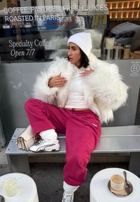 Femme en veste blanche pelucheuse, bonnet et pantalons de survêtement rouges assise sur un banc en métal à l'extérieur d'un café avec une tasse de café et une limonade sur de petites tables à proximité.