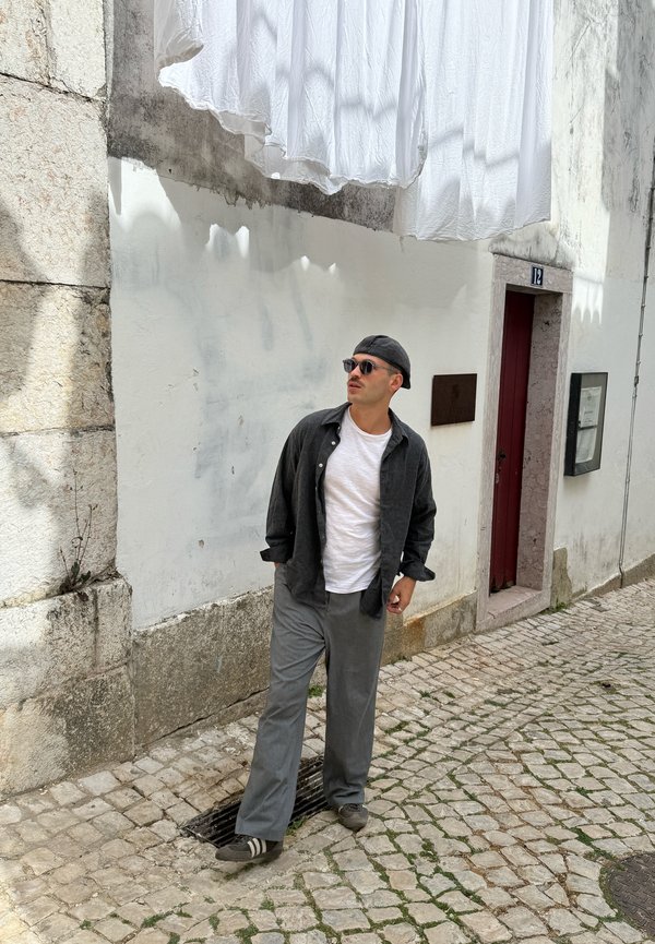 Mann mit Sonnenbrille, dunklem Hemd, weißem T-Shirt, grauer Hose und Kappe steht auf gepflasterter Straße unter hängenden weißen Laken nahe einer weißen Wand und einer roten Tür.