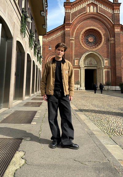 Chaqueta de lona marrón sobre un suéter negro, combinada con jeans negros holgados y zapatos negros, se encuentra de pie contra el fondo de una iglesia de ladrillo.