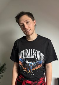 Jeune homme aux cheveux courts et foncés portant un t-shirt noir avec un graphisme coloré d'aigle et un grand texte, ainsi qu'une chemise à carreaux rouge nouée autour de la taille.